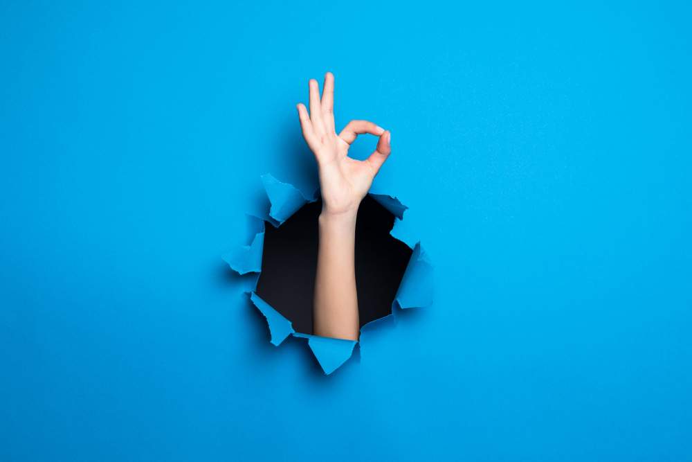 close up woman hand with okay gesture through blue hole paper wall 11zon